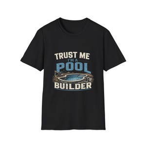 Puede incluir: Una camiseta negra de manga corta con un estampado gráfico que incluye el texto "TRUST ME I'M A POOL BUILDER" en blanco y azul. El diseño presenta una ilustración de una piscina enterrada en construcción con una excavadora cerca.