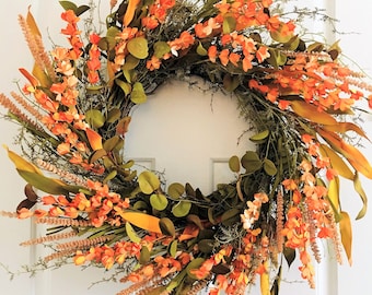 May include: A fall-themed wreath with orange and brown flowers, green leaves, and beige grasses. The wreath is on a white door with a white trim.
