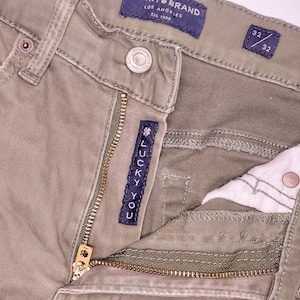 May include: A pair of olive green Lucky Brand jeans with a zipper fly and a Lucky You label. The jeans have a size tag that reads 32/32.
