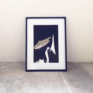 K&ouml;nnte beinhalten: Gerahmter abstrakter Kunstdruck mit einem marineblauen Rahmen. Das Kunstwerk zeigt eine goldene Pinselstrichform und wei&szlig;e und schwarze Pinselstriche auf einem dunkelblauen Hintergrund. Der Druck wird auf einem Betonboden angezeigt.