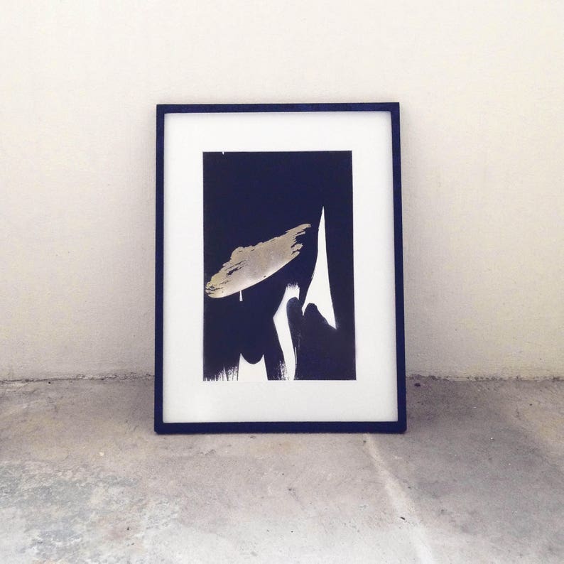 K&ouml;nnte beinhalten: Gerahmter abstrakter Kunstdruck mit einem marineblauen Rahmen. Das Kunstwerk zeigt eine goldene Pinselstrichform und wei&szlig;e und schwarze Pinselstriche auf einem dunkelblauen Hintergrund. Der Druck wird auf einem Betonboden angezeigt.