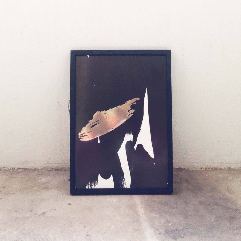 K&ouml;nnte beinhalten: Abstrakter Kunstdruck in einem schwarzen Rahmen. Das Kunstwerk zeigt einen goldenen Pinselstrich und wei&szlig;e Formen auf schwarzem Hintergrund. Die gerahmte Kunst lehnt an einer Betonoberfl&auml;che und einer wei&szlig;en Wand.