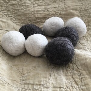 May include: Six felted wool balls, three white and three gray, arranged in a circle on a beige fabric background.