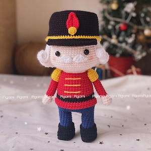 Könnte beinhalten: Eine gehäkelte Nussknackerpuppe mit roter und gelber Uniform, schwarzem Hut und weißem Schnurrbart. Die Puppe steht auf einer weißen Oberfläche mit gelben Sternen.