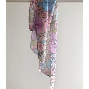 May include: A floral patterned scarf in pastel shades of pink, lilac, and blue. The scarf is suspended from a white rail, highlighting its lightweight, transparent material. The design features large, stylized flowers and foliage.