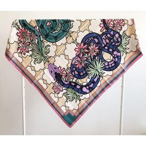 May include: Square silk scarf featuring a design of snakes and flowers. The snakes are teal and purple, with pink and green floral accents. The scarf has a cream background with a geometric pattern and a pink and green border.