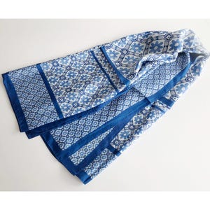 Puede incluir: Bufanda plegada con estampado azul y blanco. La bufanda presenta un diseño floral y geométrico repetitivo en tonos azules. Bordes azul oscuro enmarcan los bordes y secciones, creando contraste.