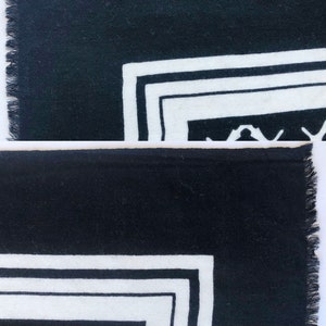 May include: Black and white patterned textile with a fringed edge. The design features a geometric border with a repeating pattern in the corner. The textile is likely a scarf or a decorative piece.