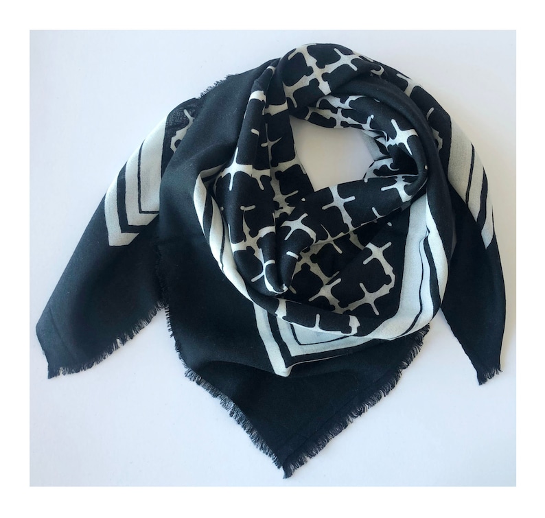 May include: A black and white square scarf with a geometric pattern. The scarf has a black background with a white geometric design in the center and white borders with a chevron pattern. The edges are fringed.