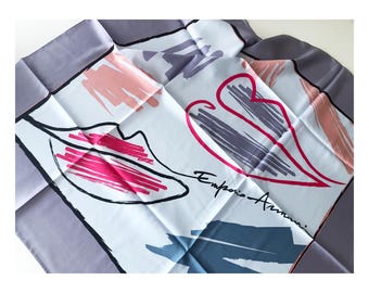 May include: A square silk scarf with a white background and a gray border. The scarf features abstract designs in pink, red, and blue, with the words "Emporio Armani" in black script.
