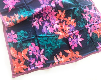 May include: A square, navy blue scarf with a vibrant floral pattern. The design features flowers in shades of pink, orange, purple, and teal. The scarf has a dark purple border.