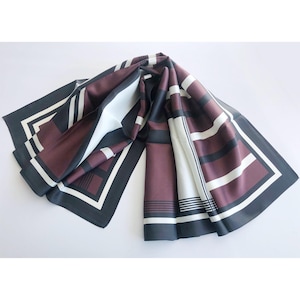 May include: A folded silk scarf with a geometric pattern. The scarf features a color palette of burgundy, black, white, and gray. The design includes stripes and rectangular shapes, with a black border. The scarf is displayed on a white surface.