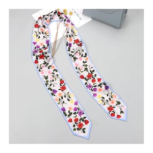 May include: A long, narrow scarf featuring a white background with a repeating pattern of red, purple, pink, and yellow roses on green stems. The scarf has a light blue border along the edges and pointed ends, creating a floral accessory.