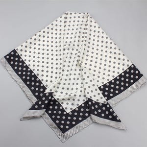 Peut inclure: Un foulard carré avec un fond blanc et des pois gris. Les bords présentent une bordure noire avec de gros pois et un motif à pois plus petit. Le foulard est plié, montrant plusieurs couches.