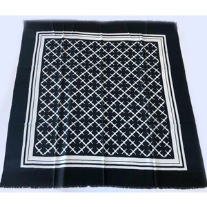 May include: A black and white square scarf with a geometric quatrefoil pattern. The scarf has a white border and fringe along the edges. The scarf is made of a soft material.
