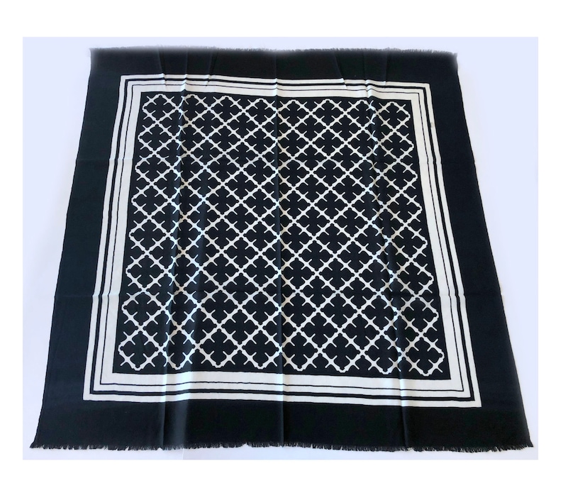 May include: A black and white square scarf with a geometric quatrefoil pattern. The scarf has a white border and fringe along the edges. The scarf is made of a soft material.