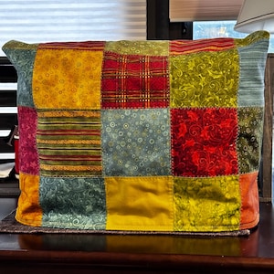 May include: A patchwork fabric item with a variety of colorful squares. The squares feature patterns like floral, plaid, and solid colors in shades of yellow, red, green, and blue. The item has a rectangular shape with rounded corners.