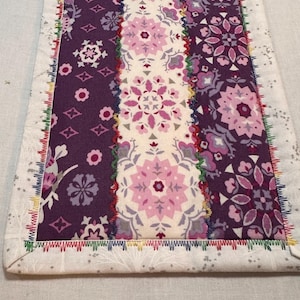 May include: A purple, pink, and white table runner with a floral pattern. The edges are finished with a decorative stitch in multiple colors.