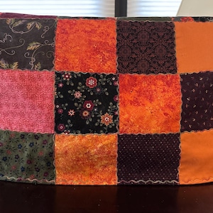 May include: A patchwork tea cozy with a variety of fabrics in shades of orange, red, pink, green, and purple. The cozy is made of squares of fabric sewn together, creating a unique and colorful design. The cozy is designed to fit over a teapot.