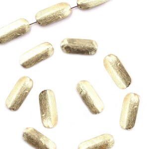 May include: A strand of gold-toned metal beads with a brushed finish. The beads are oval shaped and have a slightly irregular texture. The beads are scattered on a white background.