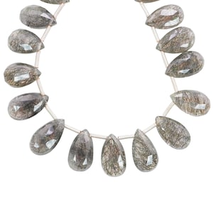 May include: A strand of faceted, teardrop-shaped gemstones. The stones are a translucent gray with dark, hair-like inclusions. The beads are strung on a thin, silver-colored cord, and are displayed against a white background. The gemstones are likely used for jewelry making.
