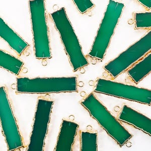 May include: A collection of rectangular green gemstone pendants with gold-coloured edging and small loops for attaching to jewellery. The pendants are scattered on a white surface, creating a visually appealing arrangement.
