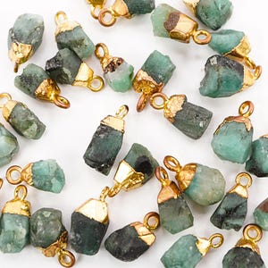 May include: A collection of emerald pendants with gold-colored caps and loops. The raw, natural emeralds vary in shades of green and are displayed against a white background. The pendants are ideal for jewelry making.
