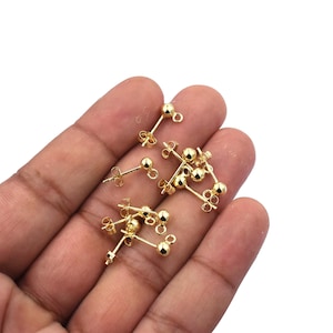 May include: A close-up of a hand holding a pile of gold-toned earring backs. The earring backs have a small ball on one end and a post with a butterfly clasp on the other end.