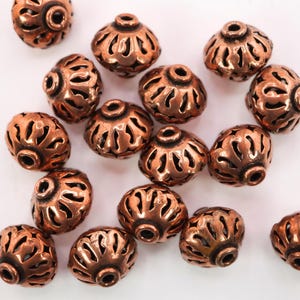 May include: A group of copper-colored metal beads with intricate cut-out designs. The beads are round and have a slightly domed shape.