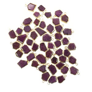 May include: A collection of irregularly shaped, deep red gemstone pendants with gold-colored metal edging. Each pendant has a small loop for attaching to jewelry. The pendants are scattered against a white background.