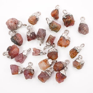 May include: A collection of raw gemstone pendants. The pendants feature rough-cut, reddish-brown stones with silver-colored metal caps and loops for hanging. The stones vary in size and shape, scattered on a white surface.