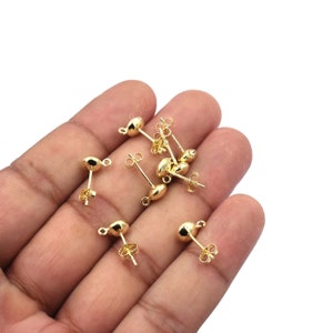 May include: A close-up of a hand holding a group of gold-toned earring posts. The posts have a small, round ball at the top and a loop at the bottom.