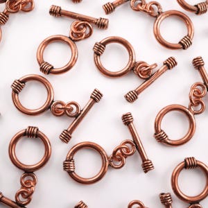 May include: Close-up of several copper-colored toggle clasps. Each clasp features a circular ring with wrapped wire detailing and a straight bar with similar wrapped wire ends. The clasps are scattered against a white background.