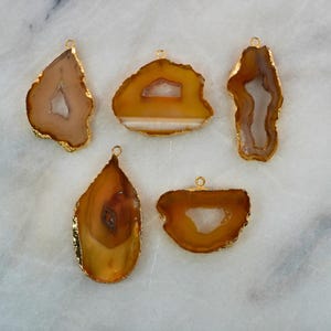 May include: Five agate pendant charms with gold-plated edges. The agate is a light brown colour with white and brown banding. The pendants are various shapes, including teardrop, oval, and irregular.
