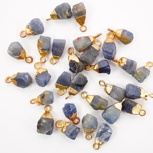 May include: A collection of raw, blue-grey gemstone pendants with gold-colored metal caps and loops. The pendants vary in size and shape, with a natural, unpolished appearance. The gold caps add a touch of elegance to the rustic stones.