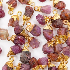 May include: Close-up of multiple raw ruby pendants with gold-colored caps and loops. The ruby stones vary in shades of pink and red, with some showing darker, more opaque areas. The gold caps add a touch of elegance to the natural gemstones.