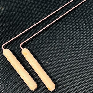 Può includere: Due strumenti in legno fatti a mano con sottili fili metallici rosa. I manici in legno sono cilindrici con un'estremità piatta. I fili metallici sono piegati ad angolo retto per collegarsi ai manici in legno.