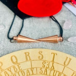 Pode incluir: Um tabuleiro Ouija de madeira com um pêndulo de cor cobre. O tabuleiro tem as letras PQRSTUVWXYZ, as palavras SIM, NÃO, TALVEZ, e os dias da semana escritos nele.