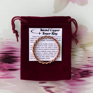 Puede incluir: Un anillo tensor retorcido de cobre descansa sobre una tarjeta blanca dentro de una bolsa de terciopelo burdeos. El anillo está hecho de alambre de cobre retorcido. La tarjeta tiene texto que describe las propiedades del anillo.