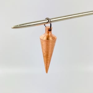 Pode incluir: Um prumo de cobre pendurado em uma haste de metal. O prumo tem a forma de um cone e tem um acabamento liso e polido.