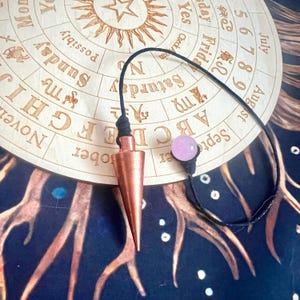 May include: A copper pendulum with a black cord and a pink crystal charm. The pendulum is hanging over a wooden board with a circular chart printed on it. The chart includes letters, numbers, and days of the week.