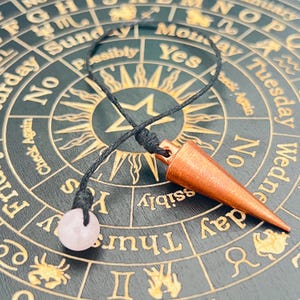Pode incluir: Um pêndulo de cor cobre com um cordão preto e um cristal rosa pendurado sobre um gráfico circular preto e dourado com os dias da semana e as palavras "Sim", "Não", "Possivelmente" e "Verificar novamente" impressas nele.