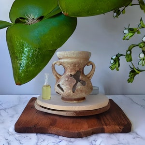 May include: A ceramic oil diffuser in the shape of a vase with a cutout design. The diffuser is brown and beige with a heart-shaped handle on each side. The diffuser is sitting on a wooden tray with a small bottle of oil and a candle.