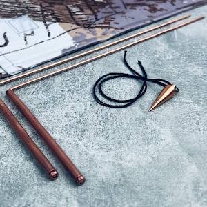 Op de afbeelding: Twee koperen wichelroeden met een puntig koperen slinger aan een zwart koord. De roeden zijn gebogen in een hoek van 90 graden.