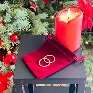 Pode incluir: Uma vela vermelha acesa e um pequeno saco de veludo vermelho com dois anéis dourados torcidos estão sobre uma superfície preta. O fundo apresenta uma árvore de Natal com enfeites vermelhos e uma flor de Natal.