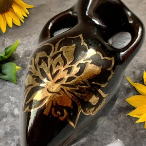May include: A black ceramic vase with a gold floral design. The vase has a unique shape and is decorated with a stylised flower.