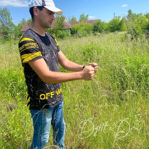 May include: A person wearing a black and white t-shirt with yellow stripes and the text "OFF-WHITE" printed on it. The person is wearing a white baseball cap and blue jeans. The person is standing in a field of tall grass.