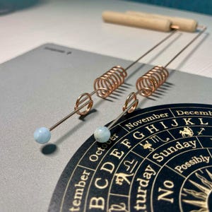 Könnte beinhalten: Zwei kupferfarbene Wünschelruten mit hellblauen Edelsteinspitzen und spiralförmigen Akzenten. Ein Holzstab befindet sich im Hintergrund. Die Ruten liegen auf einer grauen Oberfläche mit einem schwarzen und goldenen Tierkreisrad.