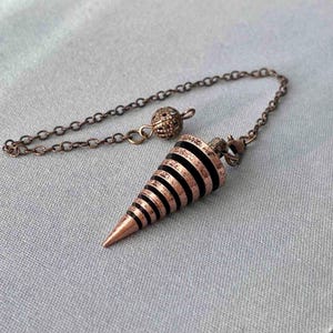May include: A copper-toned pendulum with a conical shape and black bands. It is connected to a chain and a decorative ball. The design is vintage, and the item is likely used for divination or spiritual practices.