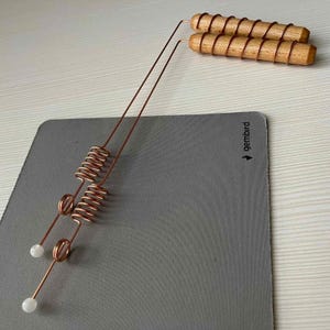 May include: Two copper dowsing rods with wooden handles wrapped in copper wire. The rods have a coiled spring design and a white, spherical tip. The rods are on a gray mousepad.